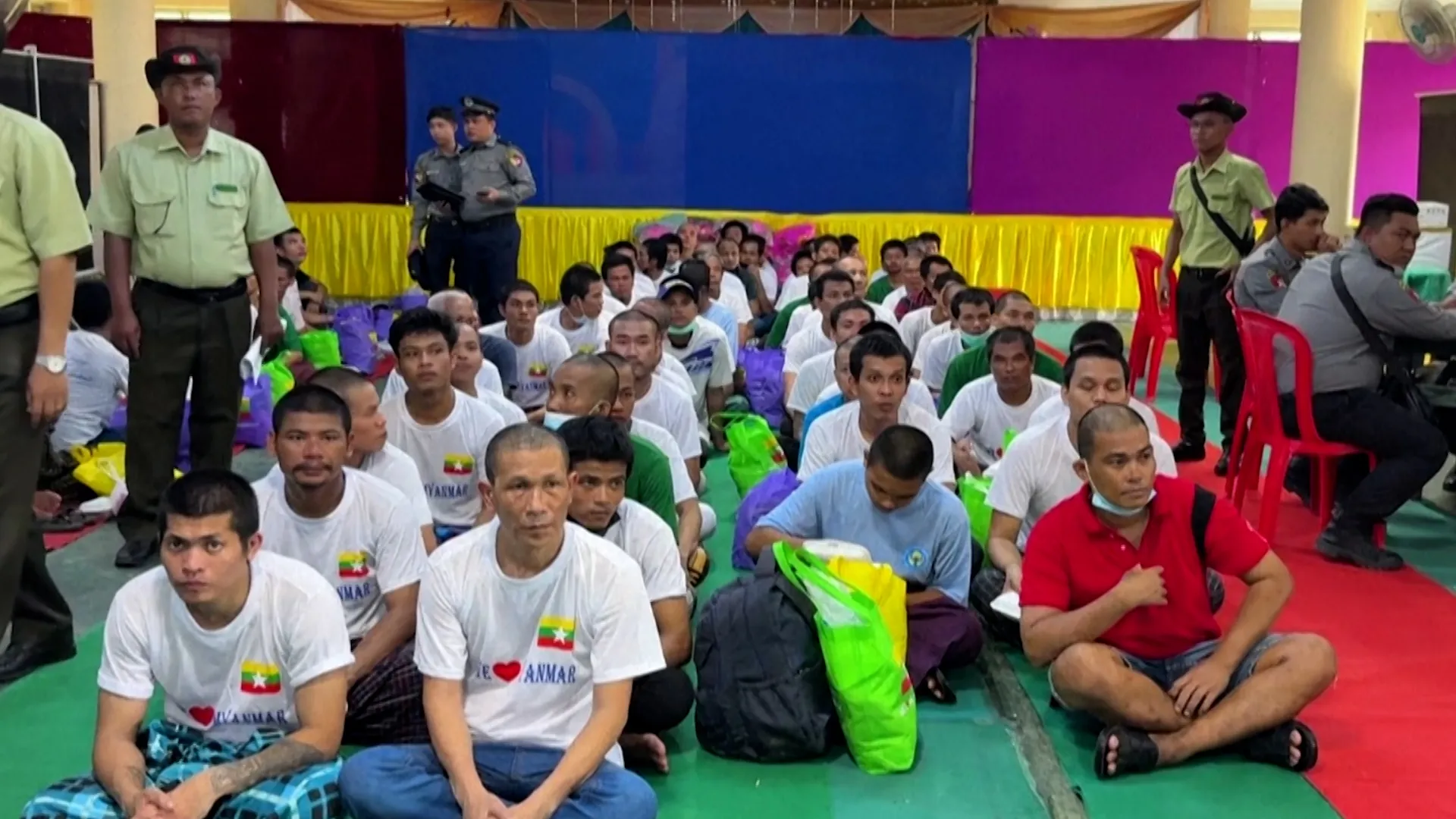 Seated men, many wearing white Myanmar flag shirts, gather indoors as uniformed officers stand nearby.
