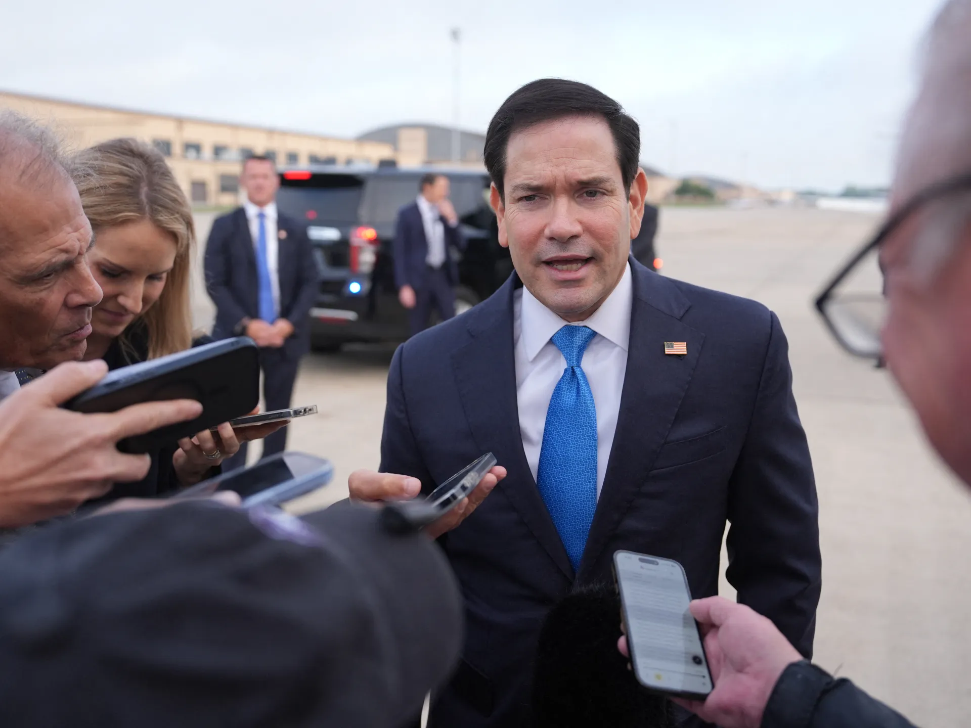 Man in a navy suit and blue tie speaks with reporters holding phones and recording devices.
