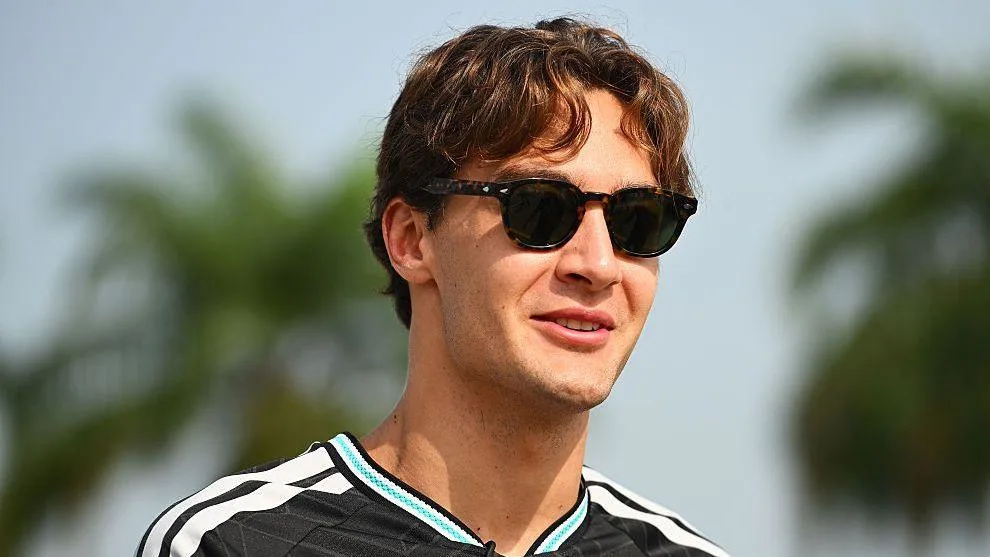 A young man in dark sunglasses and a black-and-white shirt stands outdoors against a blurred background.