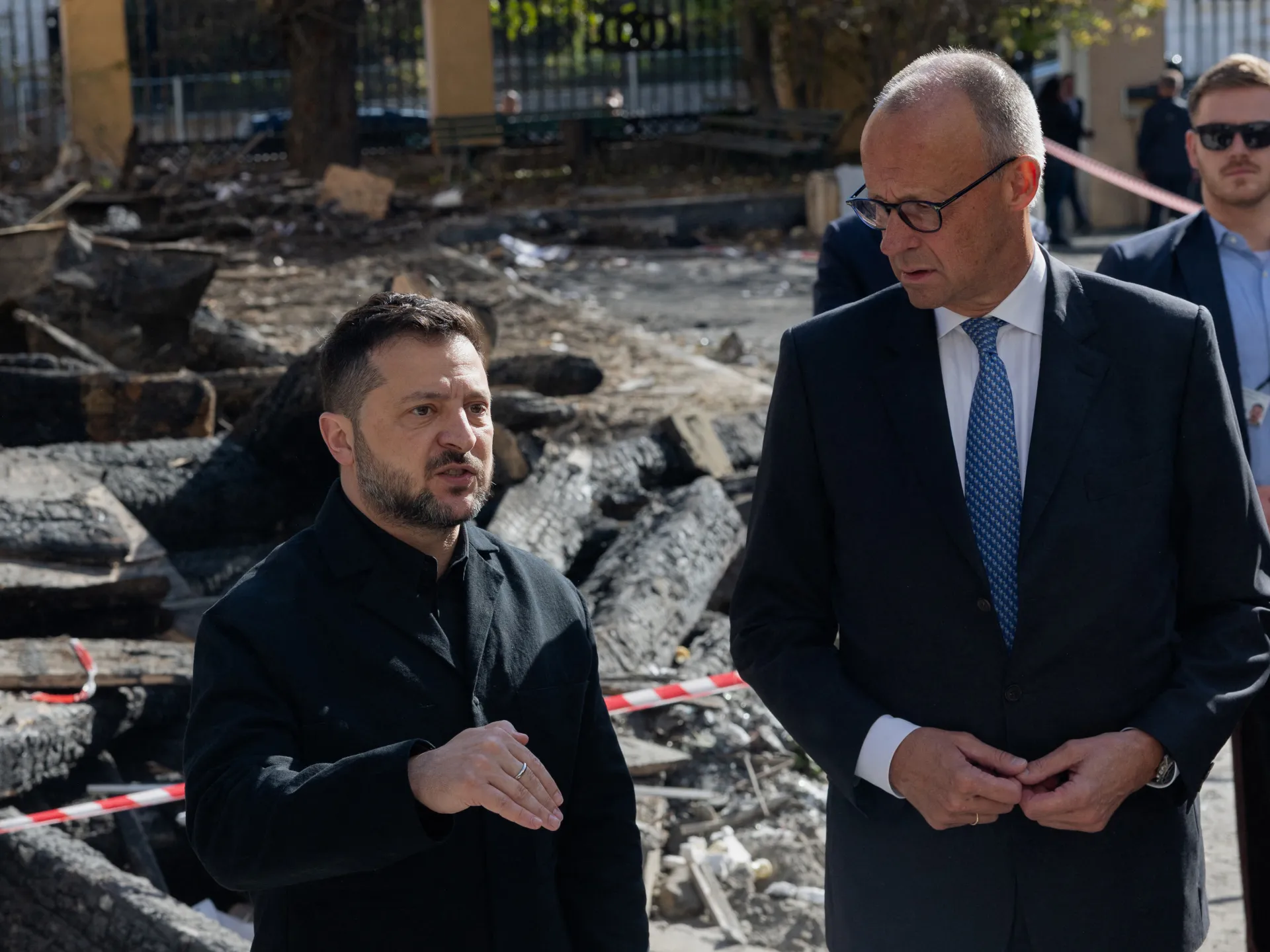 Two men in dark coats stand among rubble and damaged structures, with security personnel behind them.