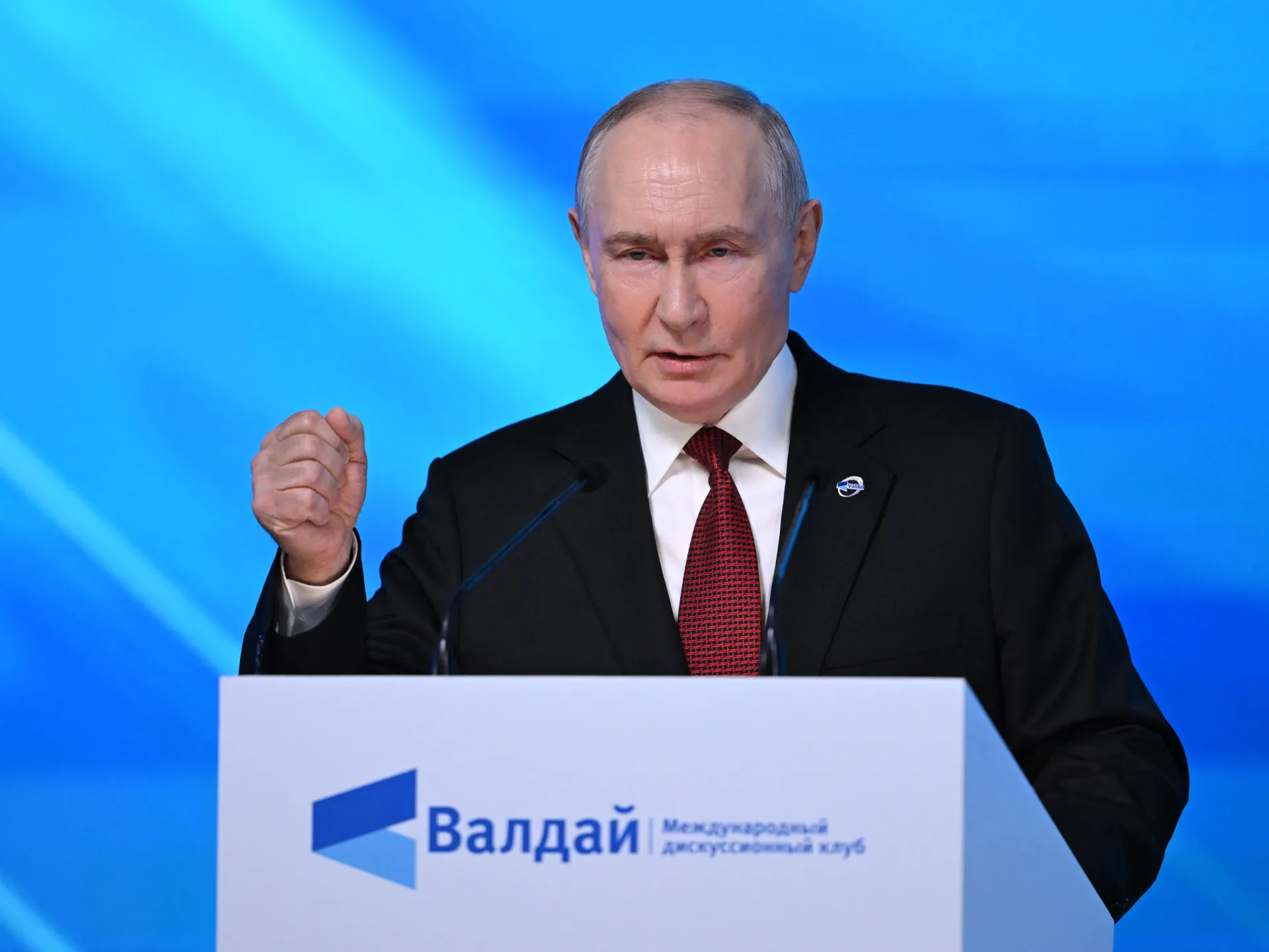 A suited man gestures behind a lectern with Russian text against a bright blue backdrop.
