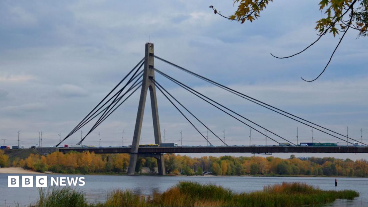 A cable-stayed bridge crosses a river, with autumn trees along the banks beneath a cloudy sky.
