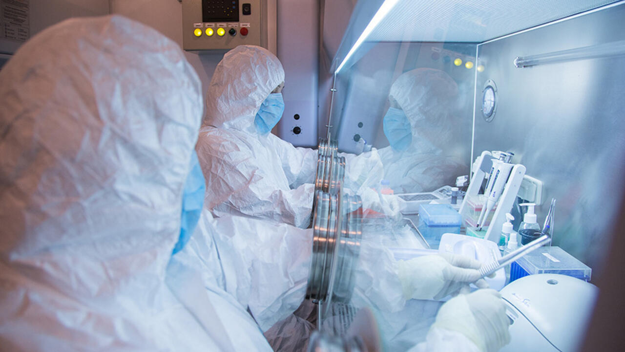 People in protective suits work with laboratory equipment behind a glass enclosure.