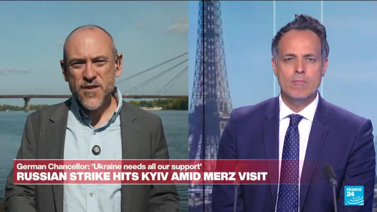 Split-screen broadcast shows two men speaking, with a river bridge behind the left speaker and a tower behind the right.