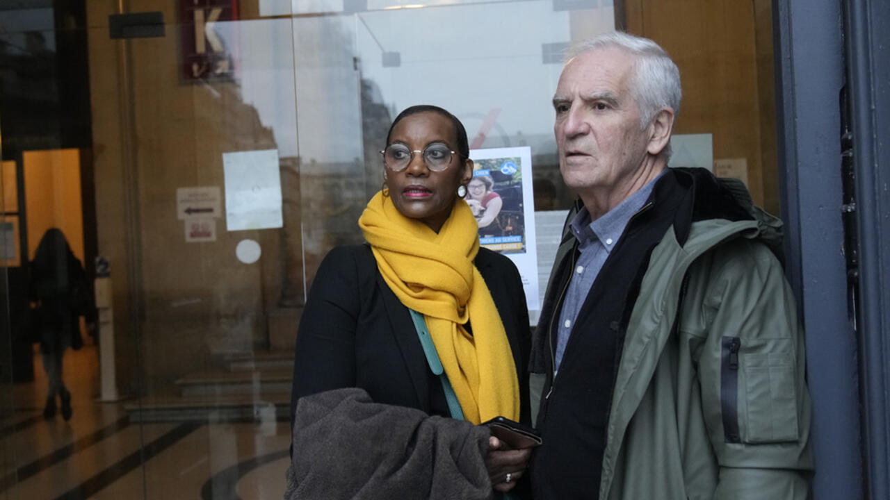 A woman in a yellow scarf and an older man stand outside a glass-fronted building.
