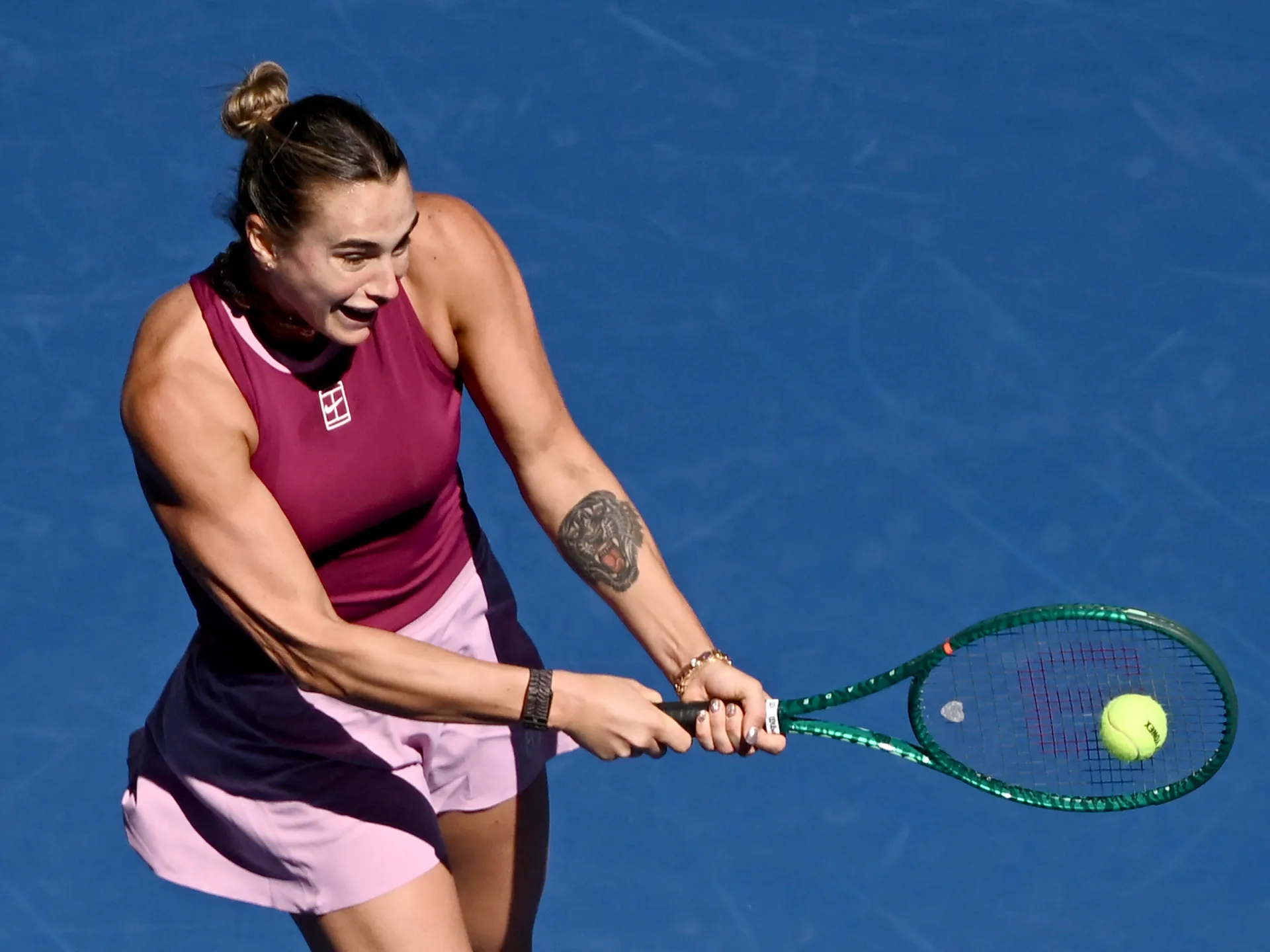 Tennis player in a burgundy and pink outfit swings a racket toward a ball.