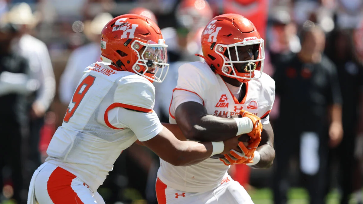 Two Sam Houston football players in white uniforms and orange helmets execute a handoff during a game.