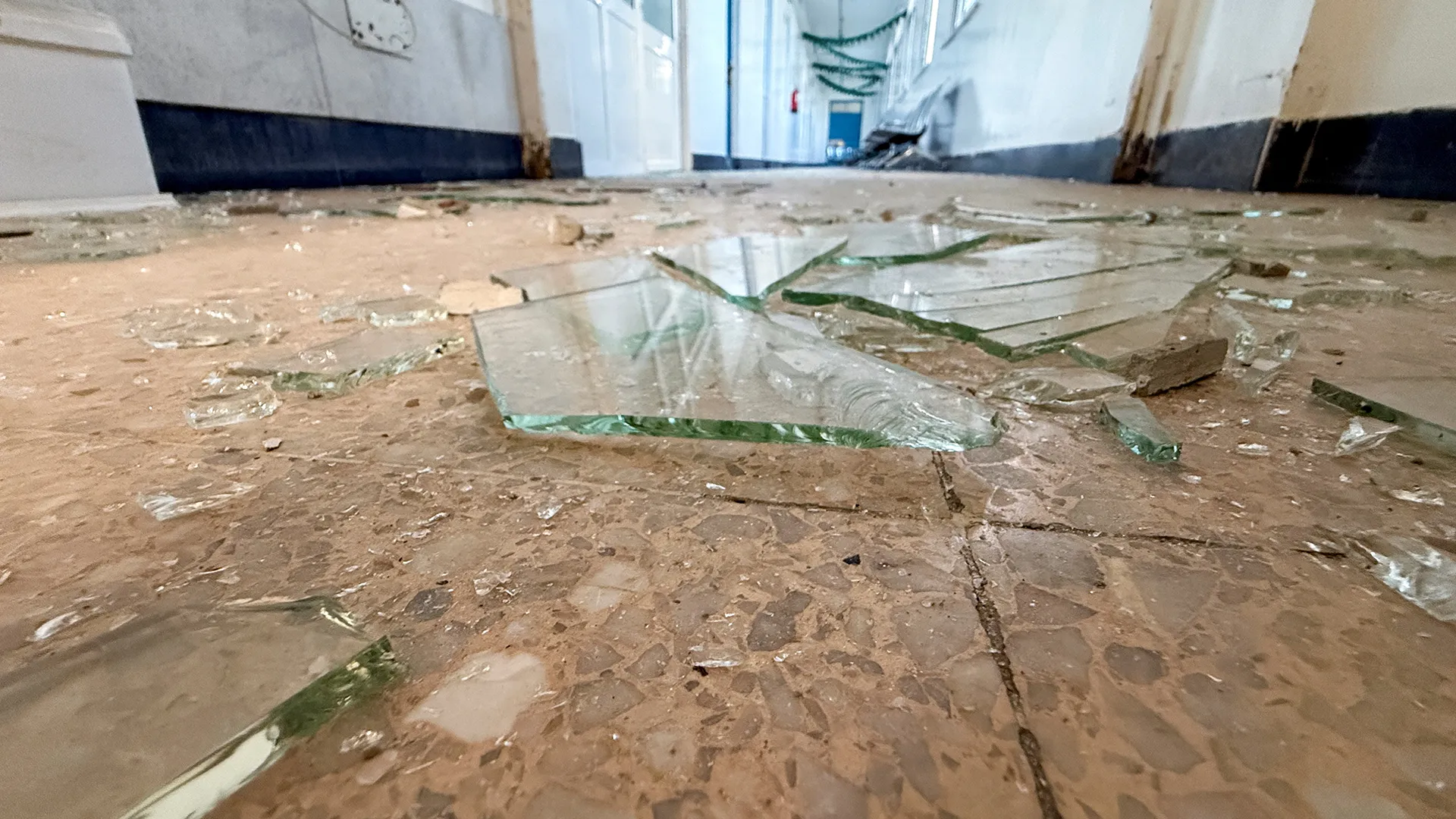 Broken glass and debris scattered across a tiled hallway floor, with damaged walls in the background.