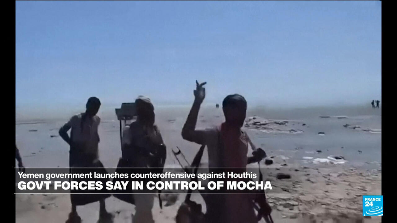 Armed men stand on a barren shoreline, one raising his hand toward the camera.