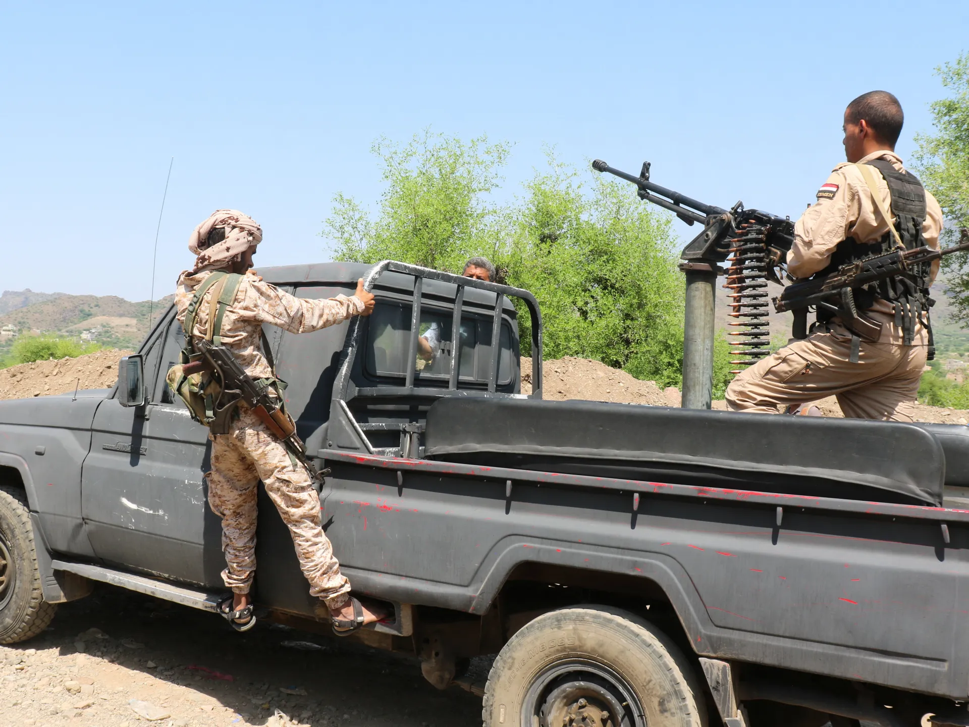Two armed men in military clothing stand beside a pickup truck fitted with a mounted machine gun.