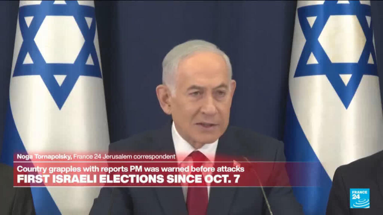 A gray-haired man in a dark suit and red tie speaks between two Israeli flags, with a news banner below.