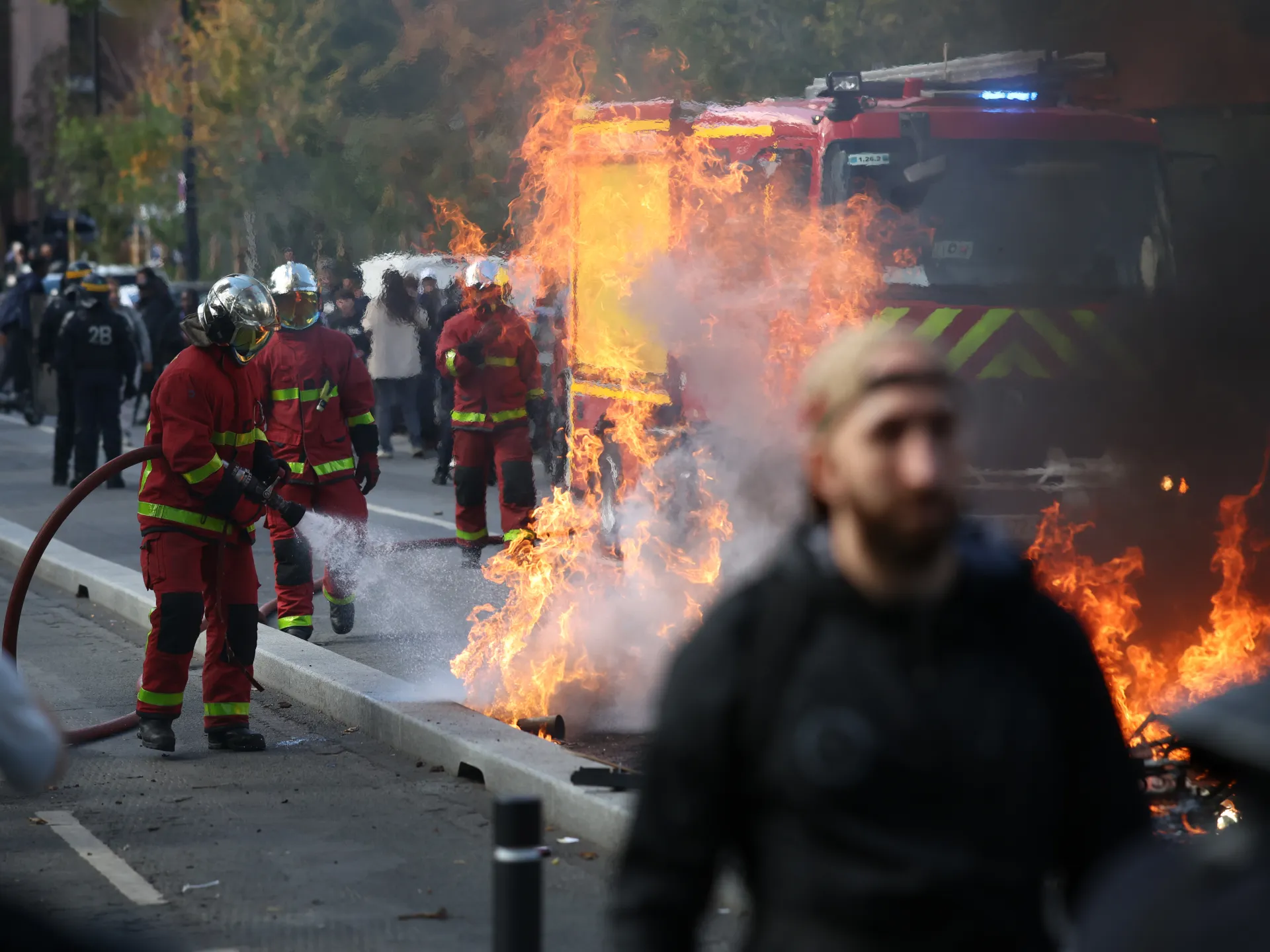 Firefighters spray water beside a burning vehicle as flames and smoke rise near a blurred passerby.