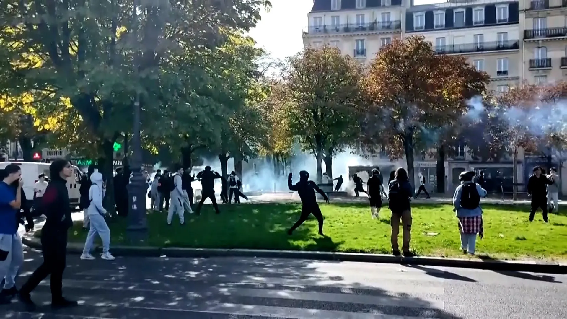 People gather on a grassy square as smoke drifts through trees beside city buildings.