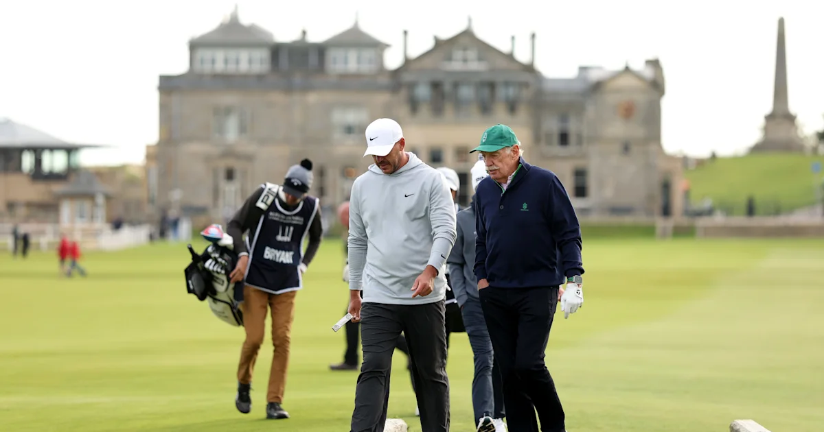 Two golfers walk across a course, followed by a caddie carrying clubs, with a large building behind them.