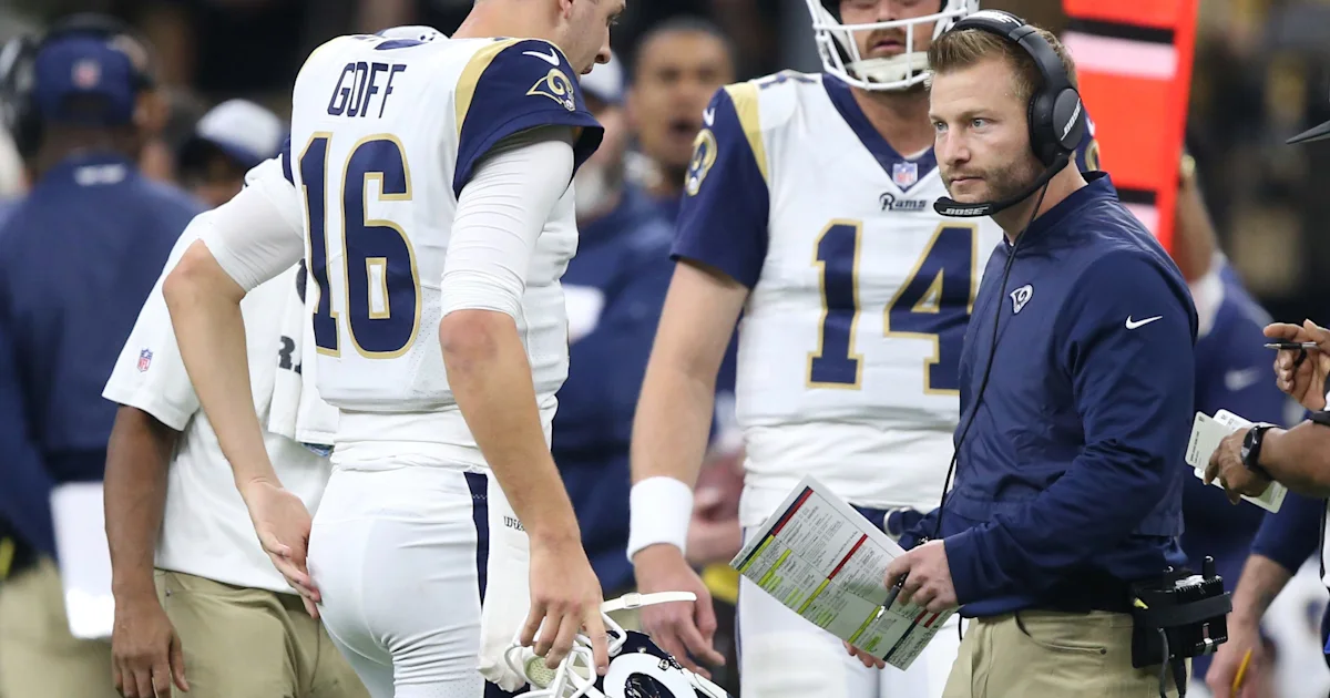 Rams coach in a headset holds a play sheet beside two players in white jerseys.