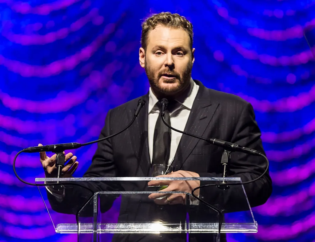 A bearded man in a dark suit speaks at a clear lectern against a blue and purple stage backdrop.
