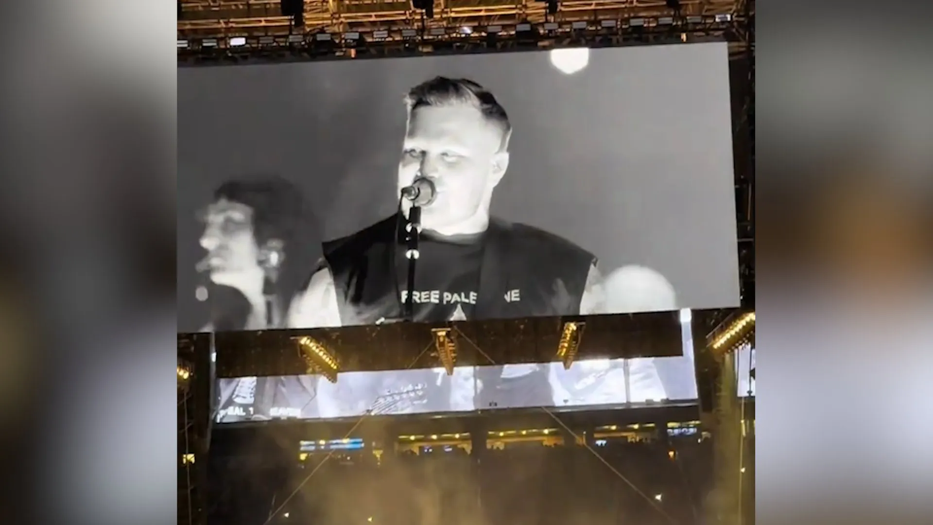 A performer sings into a microphone on a large stadium screen, wearing a dark shirt with partial white lettering.