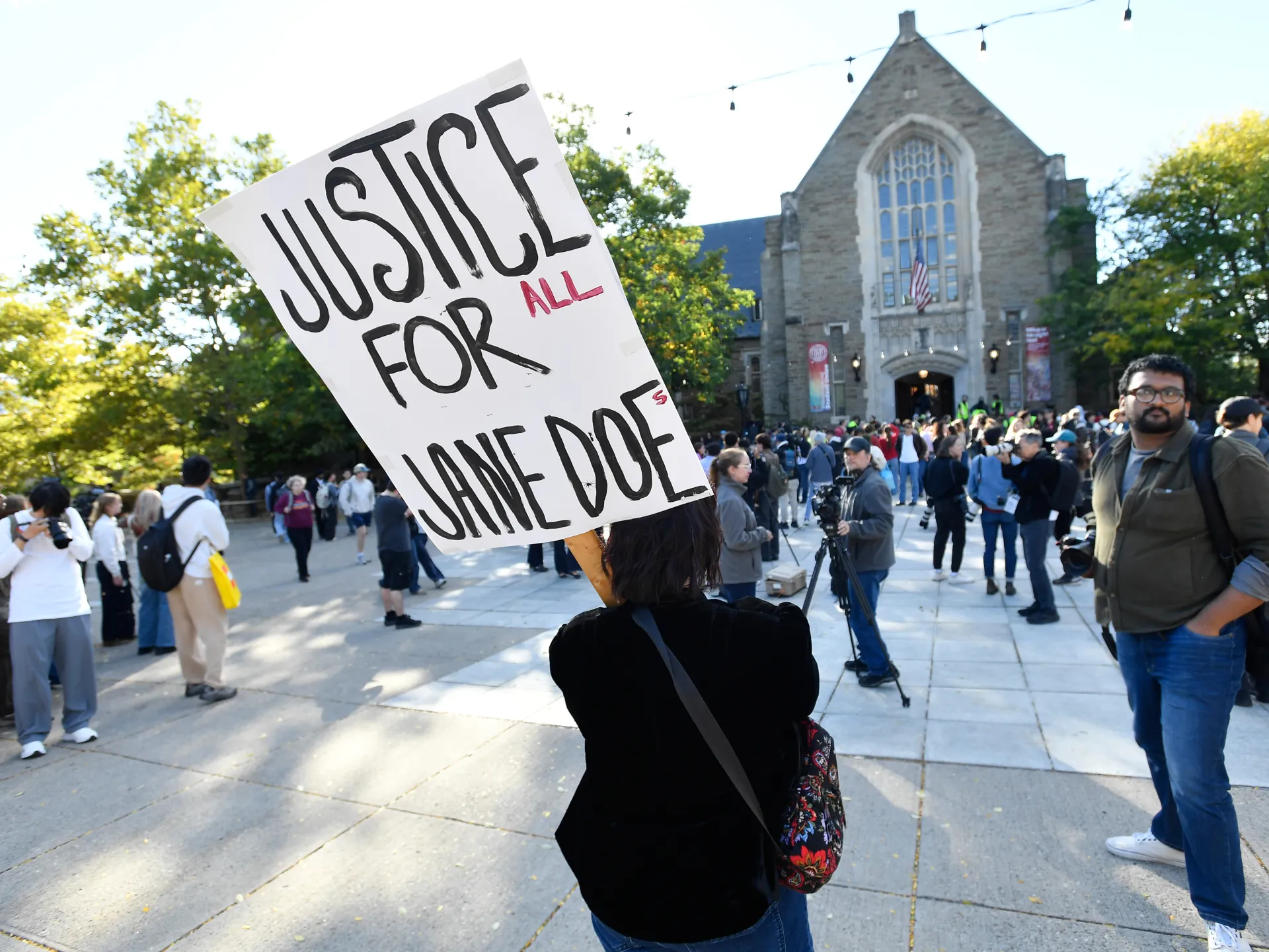 A protester holds a “Justice for all” sign outside a stone building as a crowd gathers.