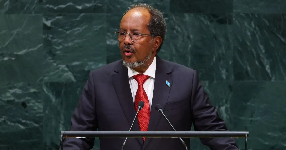 A suited man wearing glasses and a red tie speaks at a lectern before a green marble wall.
