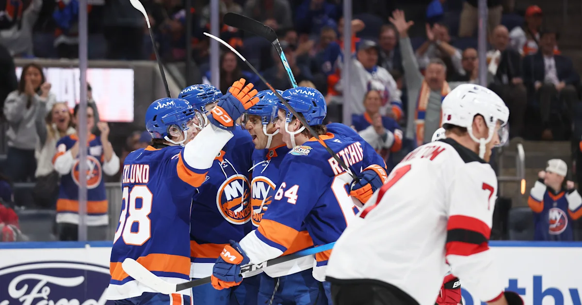 Islanders players in blue celebrate together as a Devils player in white skates past.