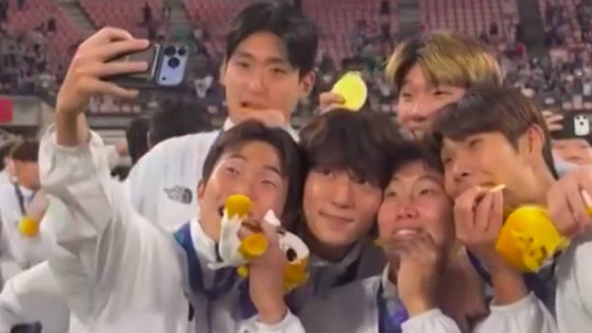 Several young men in white jackets crowd together for a selfie, holding yellow plush items in a stadium.