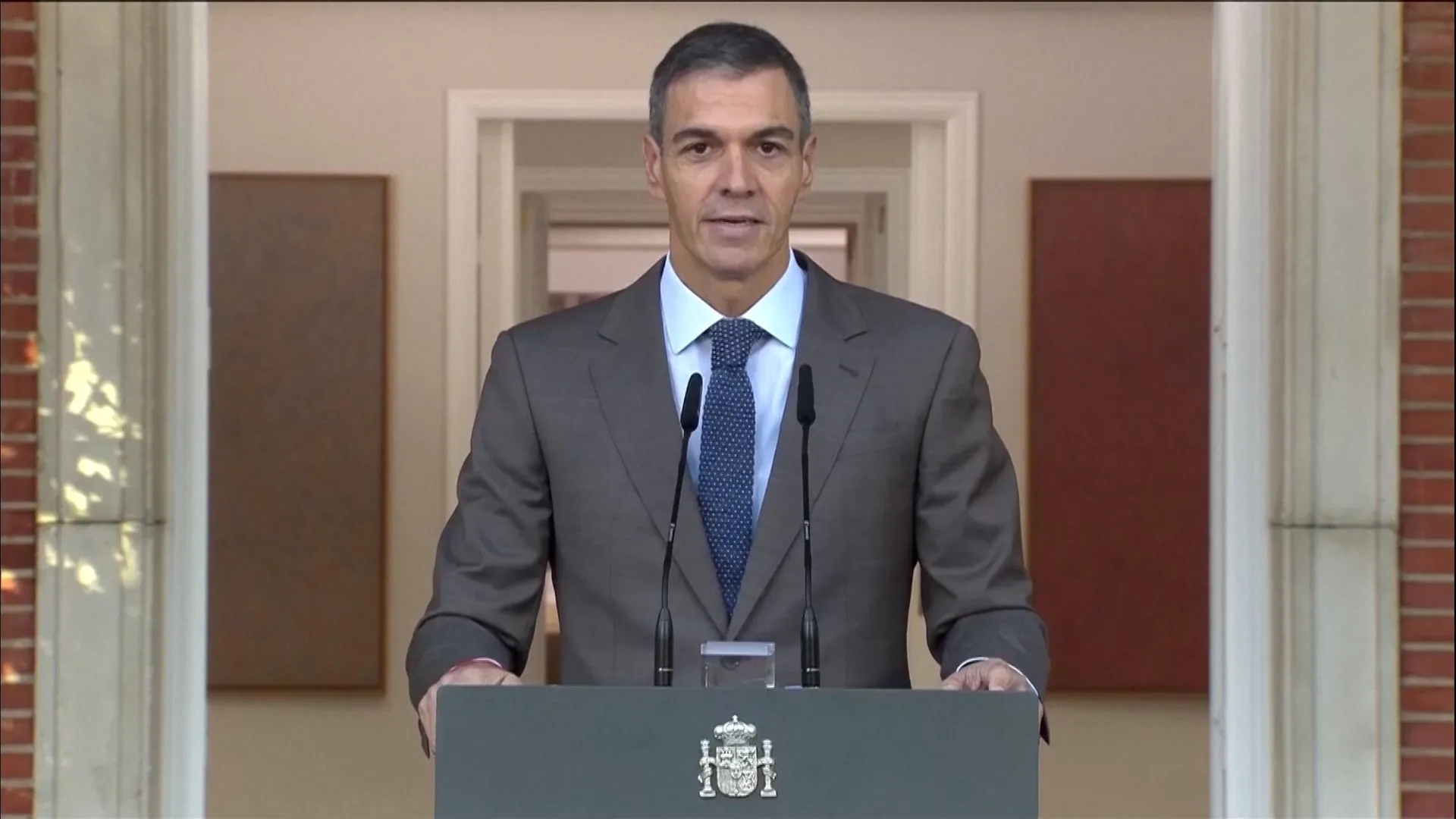 A suited man stands behind a lectern with two microphones in a formal doorway.