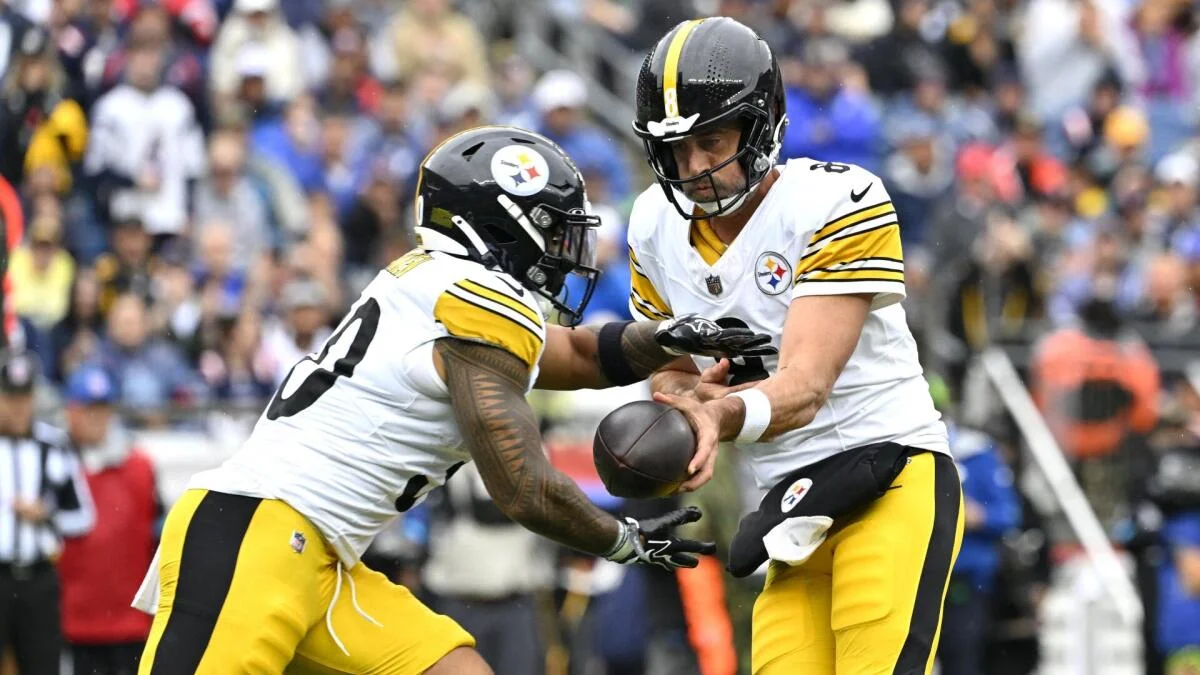 Two Pittsburgh Steelers players handle a football during a game, with a blurred crowd in the background.