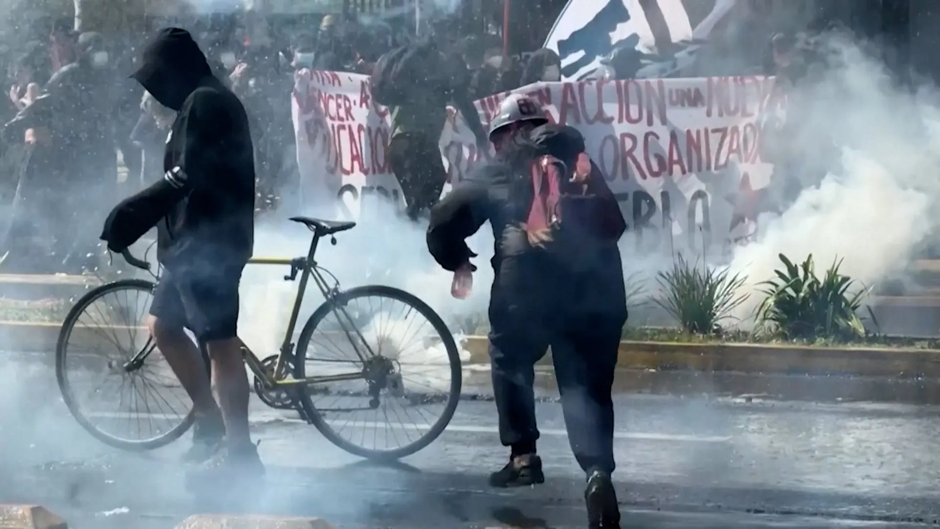 Smoke surrounds protesters, including a hooded person beside a bicycle and another person in dark clothing.