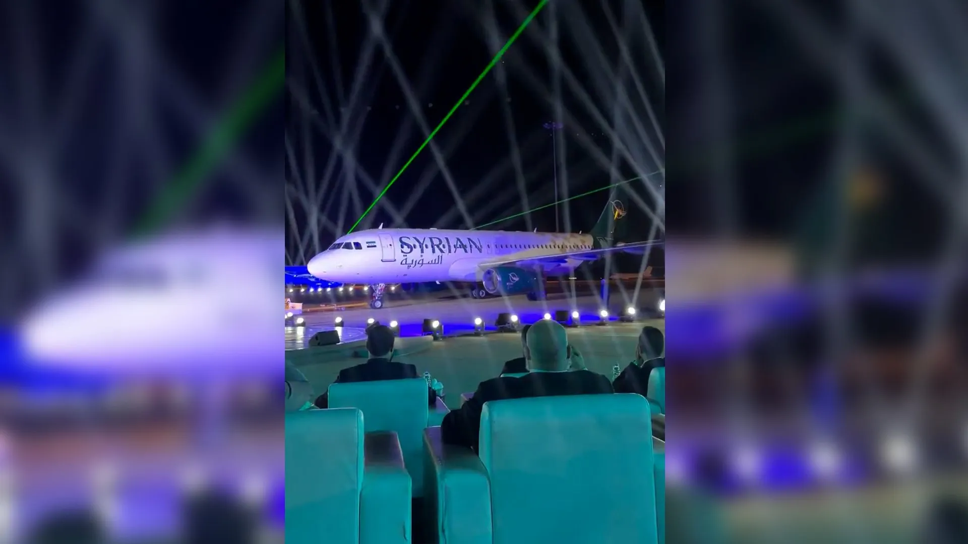 Syrian Airlines passenger jet sits on a runway amid green and white laser beams and seated spectators.