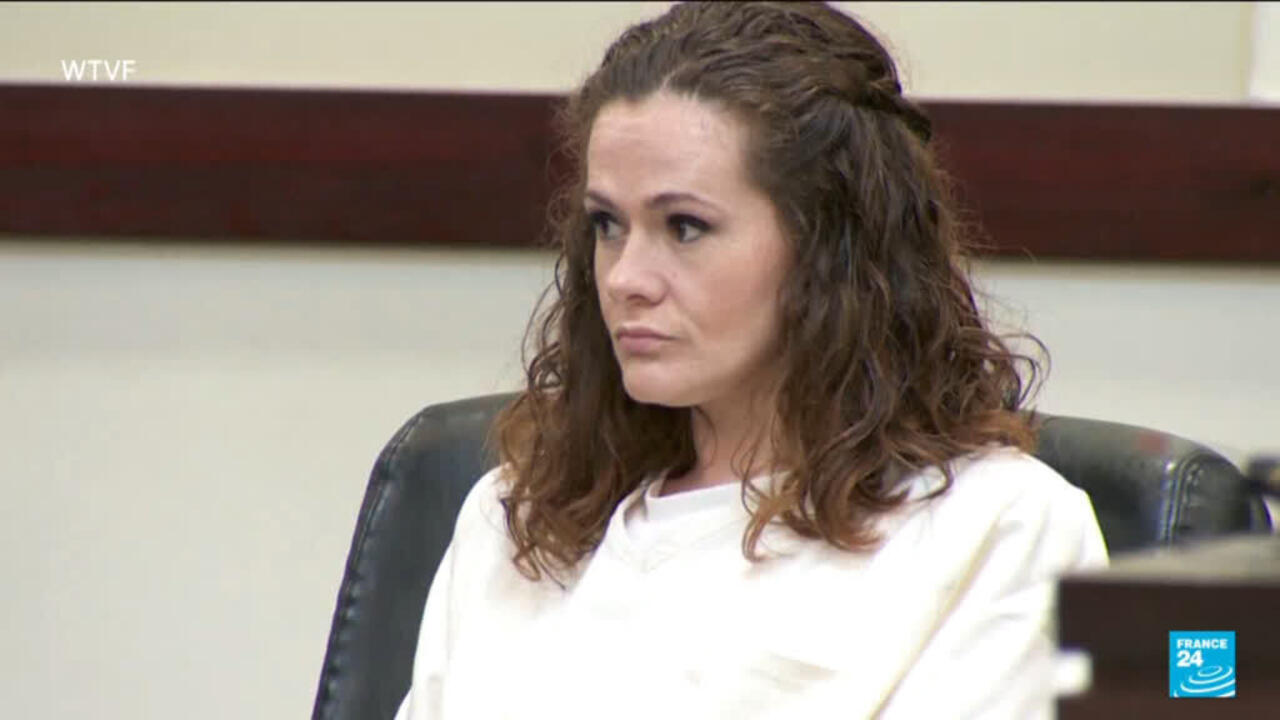 A woman in a white top sits in a black chair in a courtroom.