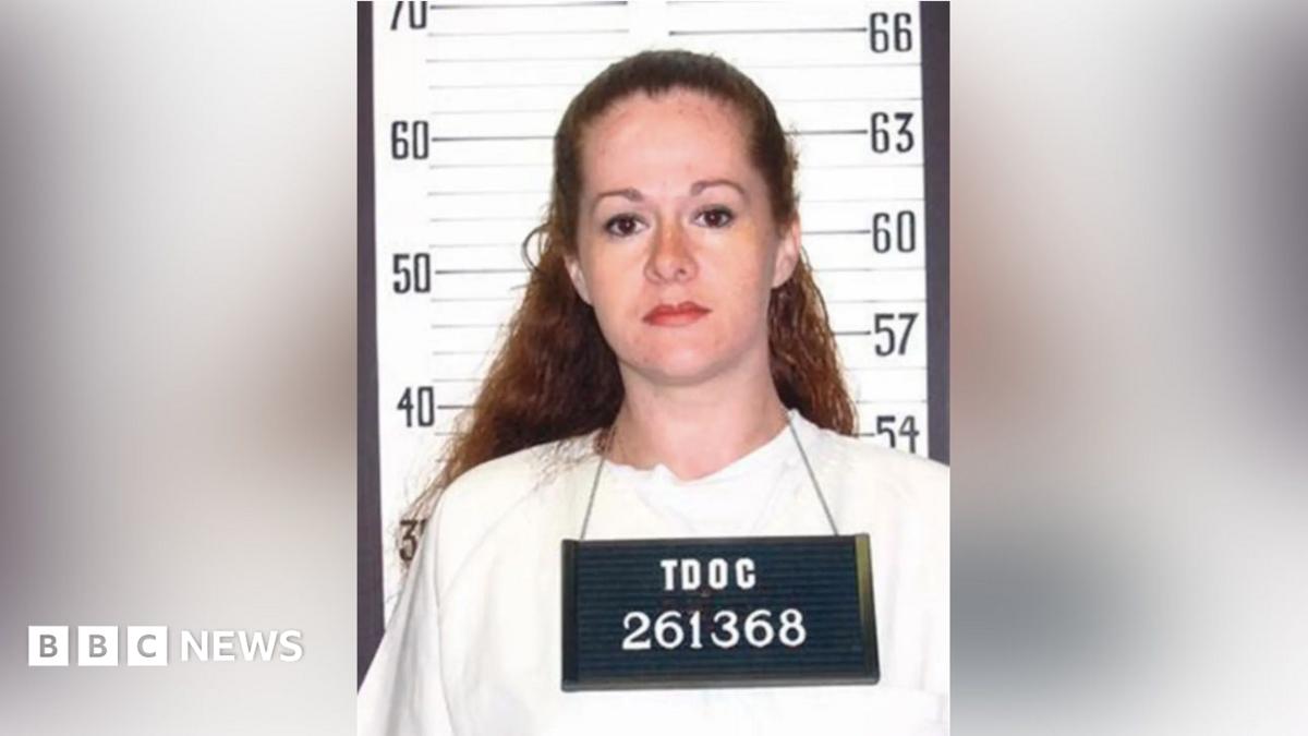 Woman with reddish hair in a white shirt holds a booking board before a height chart.