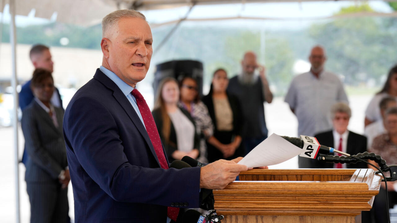 A suited man reads from a paper at a wooden lectern as microphones point toward him.