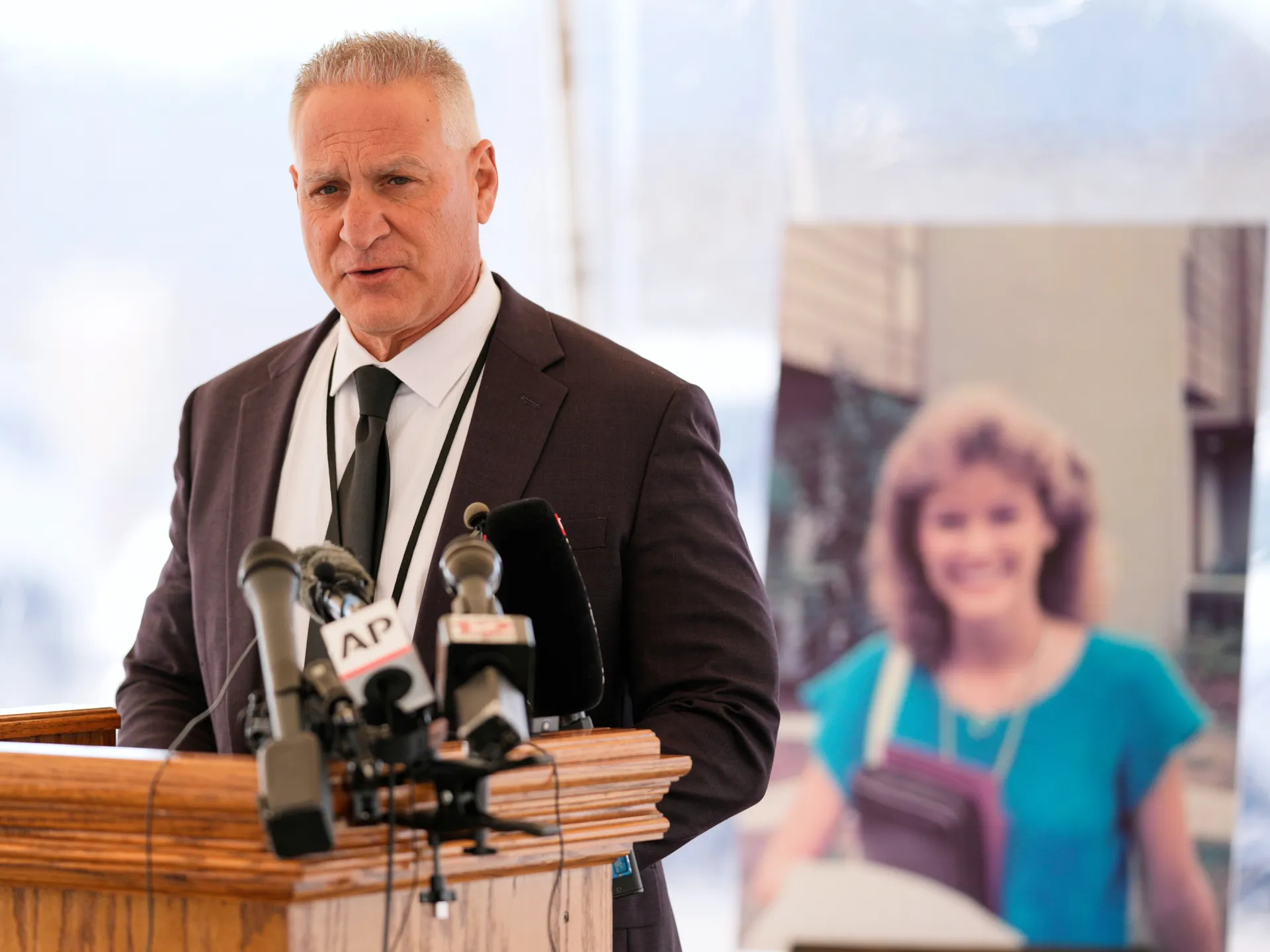 Man in a dark suit speaks at a wooden lectern crowded with microphones; a woman appears blurred behind him.