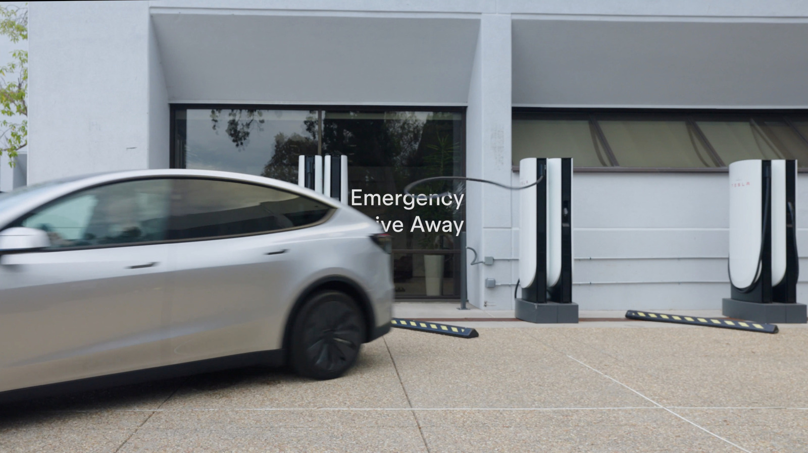 A white Tesla drives past several Supercharger stalls outside a modern building.