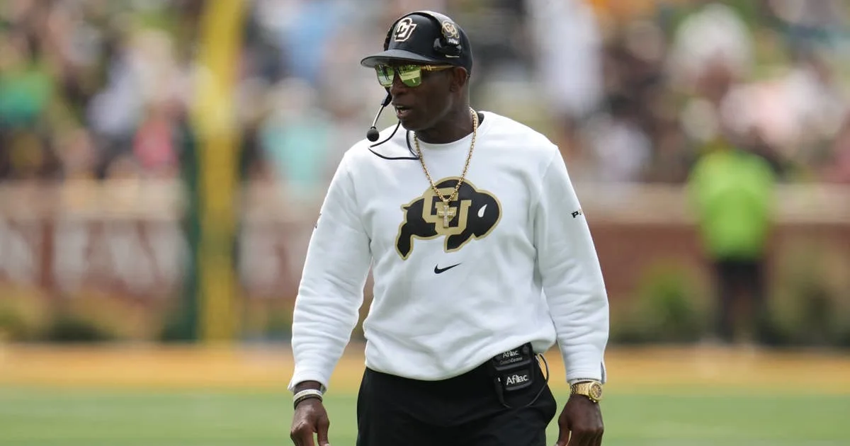 A football coach in a white Colorado sweatshirt, black cap, sunglasses, and headset stands on a field.