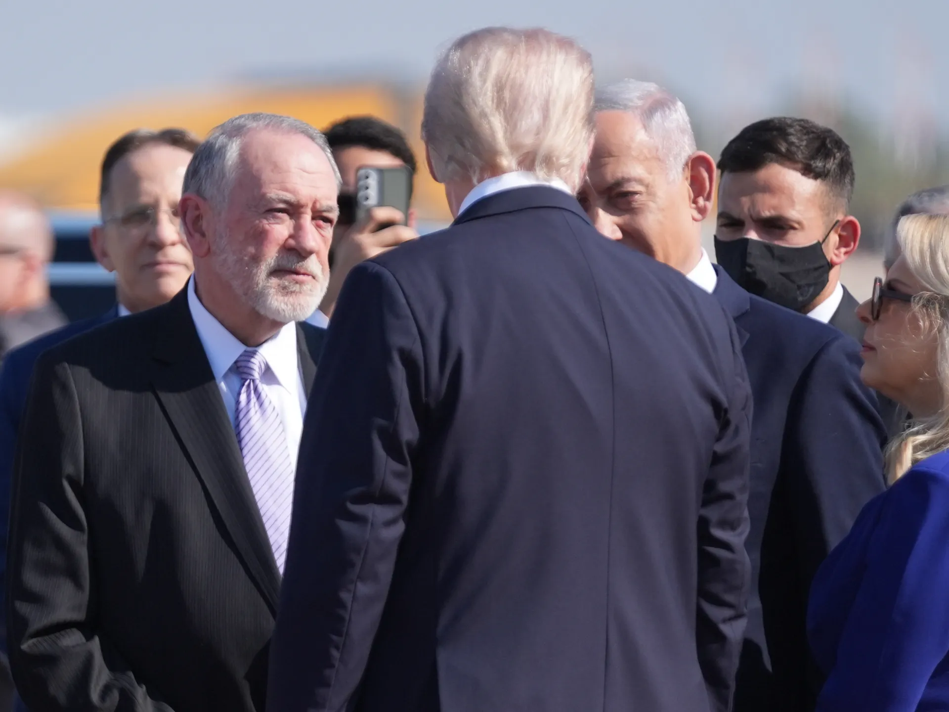 Donald Trump, seen from behind, faces several suited men, including a gray-haired bearded man and another resembling Benjamin Netanyahu.