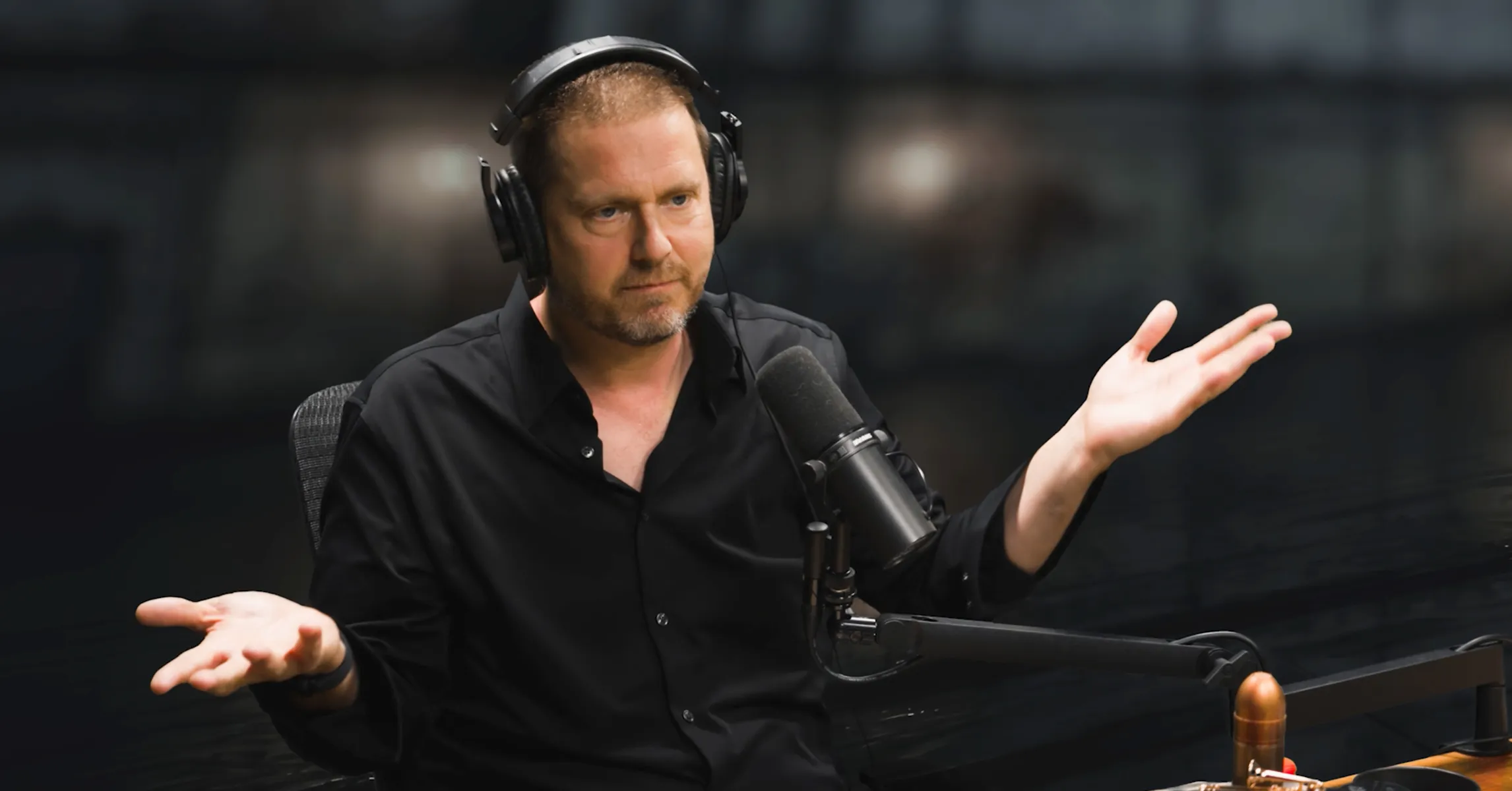 A man wearing headphones gestures with both hands beside a microphone in a dark recording studio.