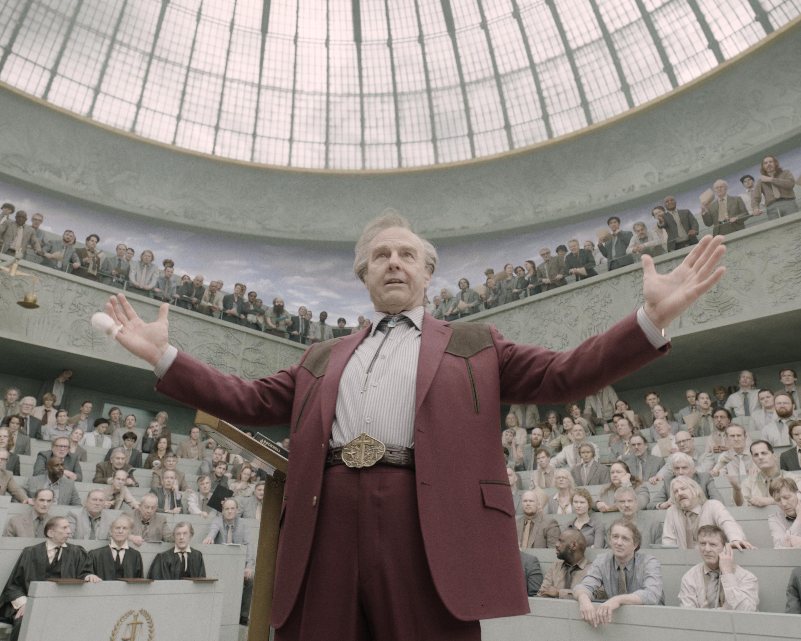Man in a burgundy suit stands with arms outstretched before a crowded, domed auditorium.