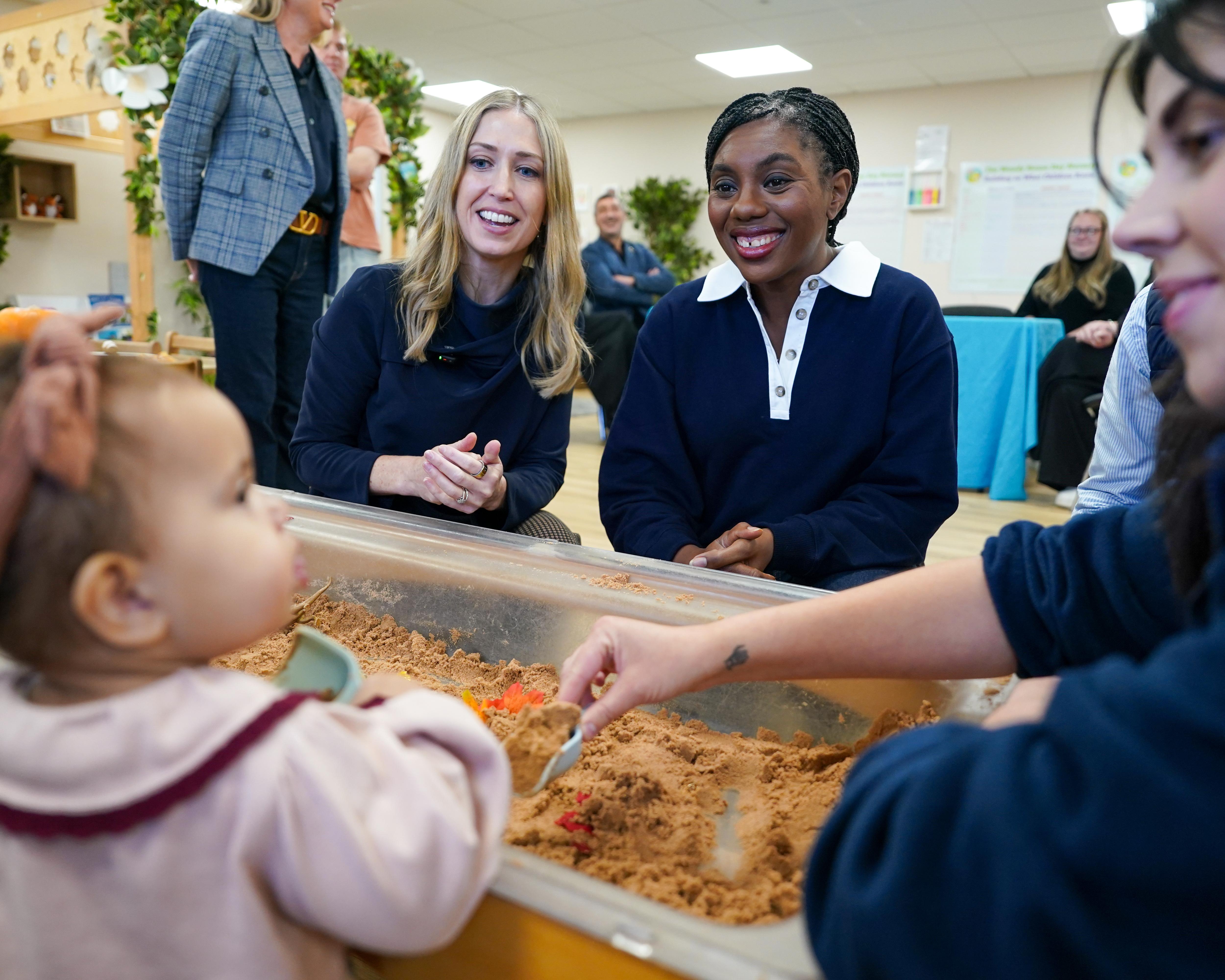 Two women sit beside a child playing with sand in a classroom activity area.