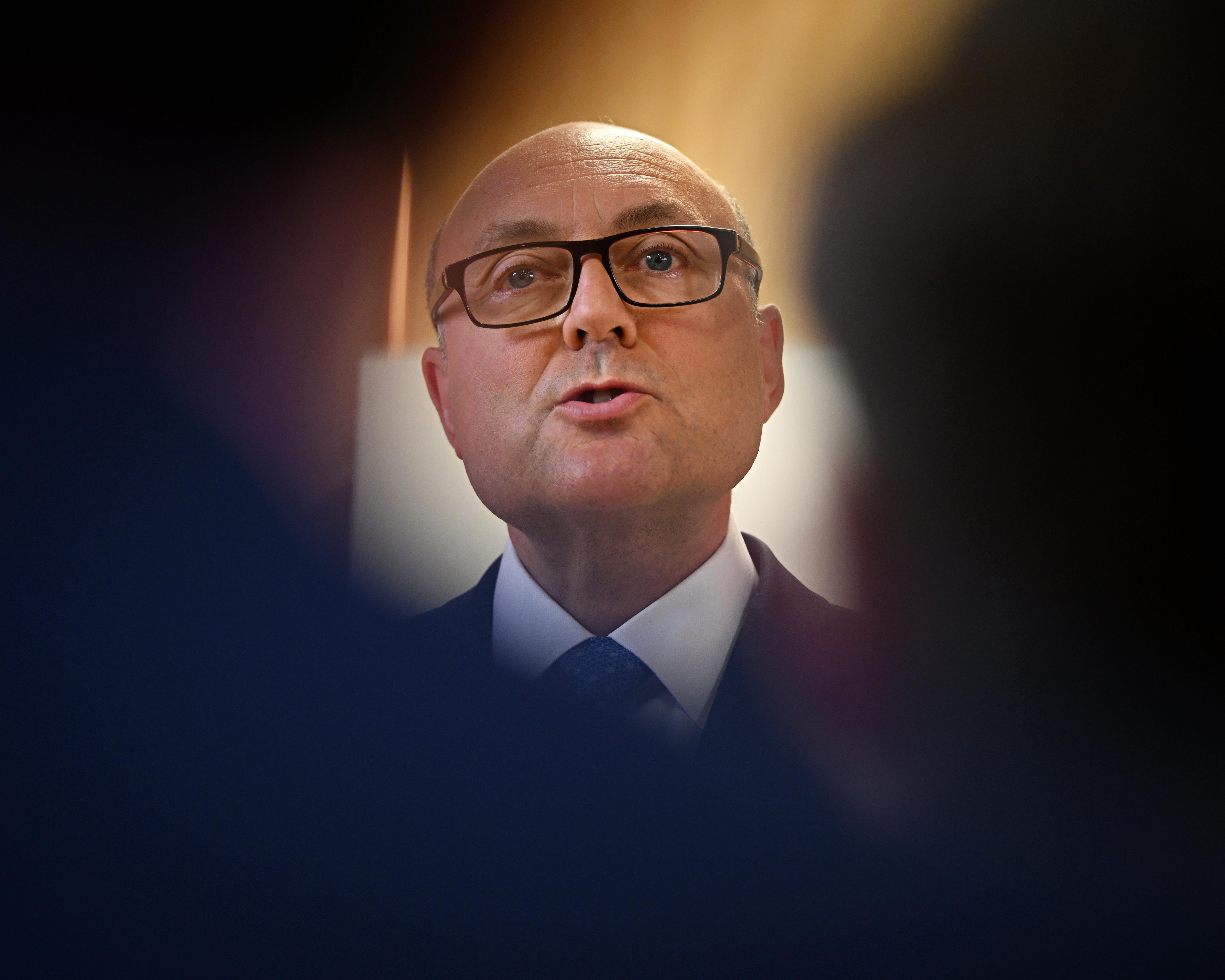 Bald man wearing glasses, a dark suit, white shirt and blue tie, photographed under warm lighting.