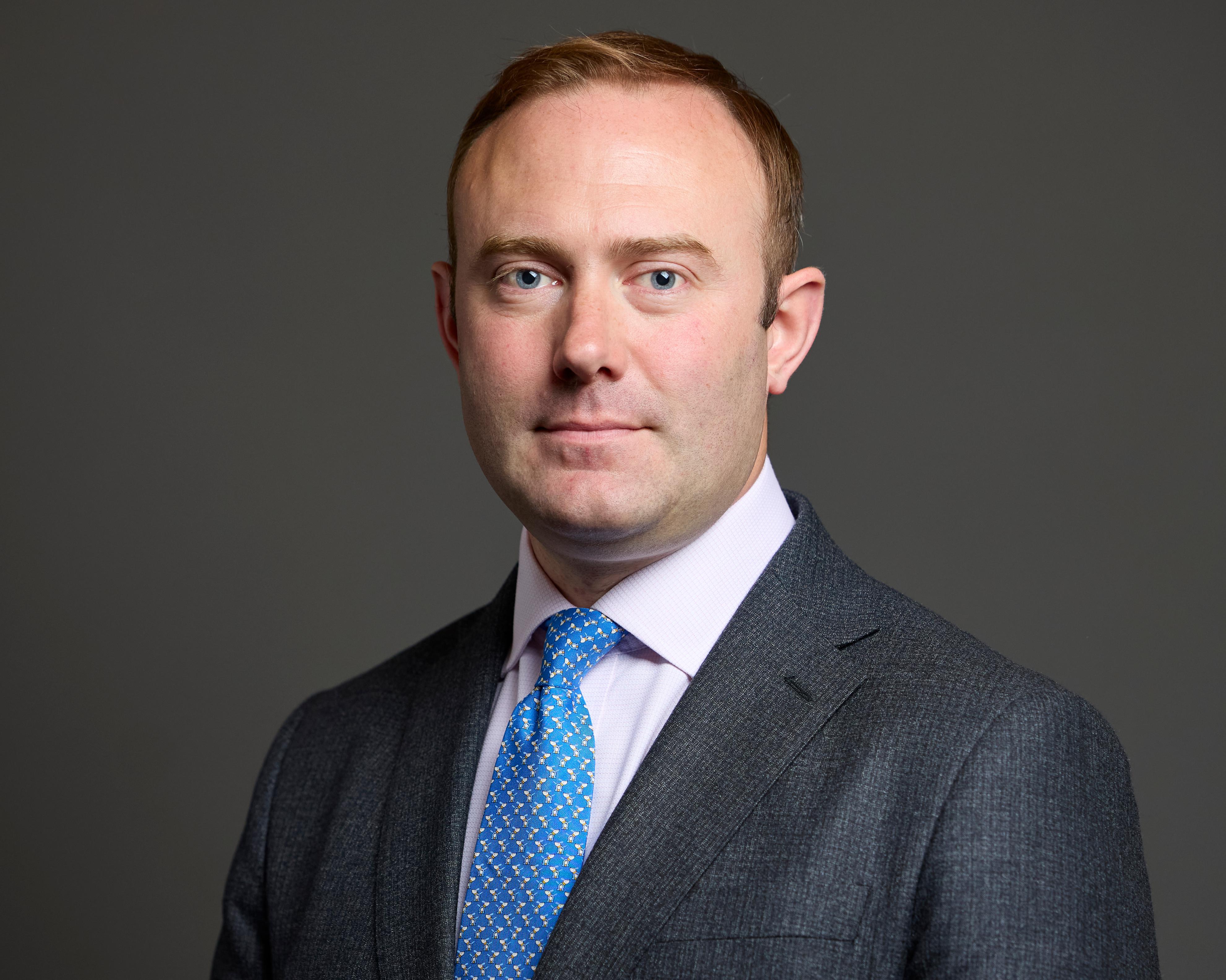 A man in a dark suit and blue patterned tie poses against a plain gray background.