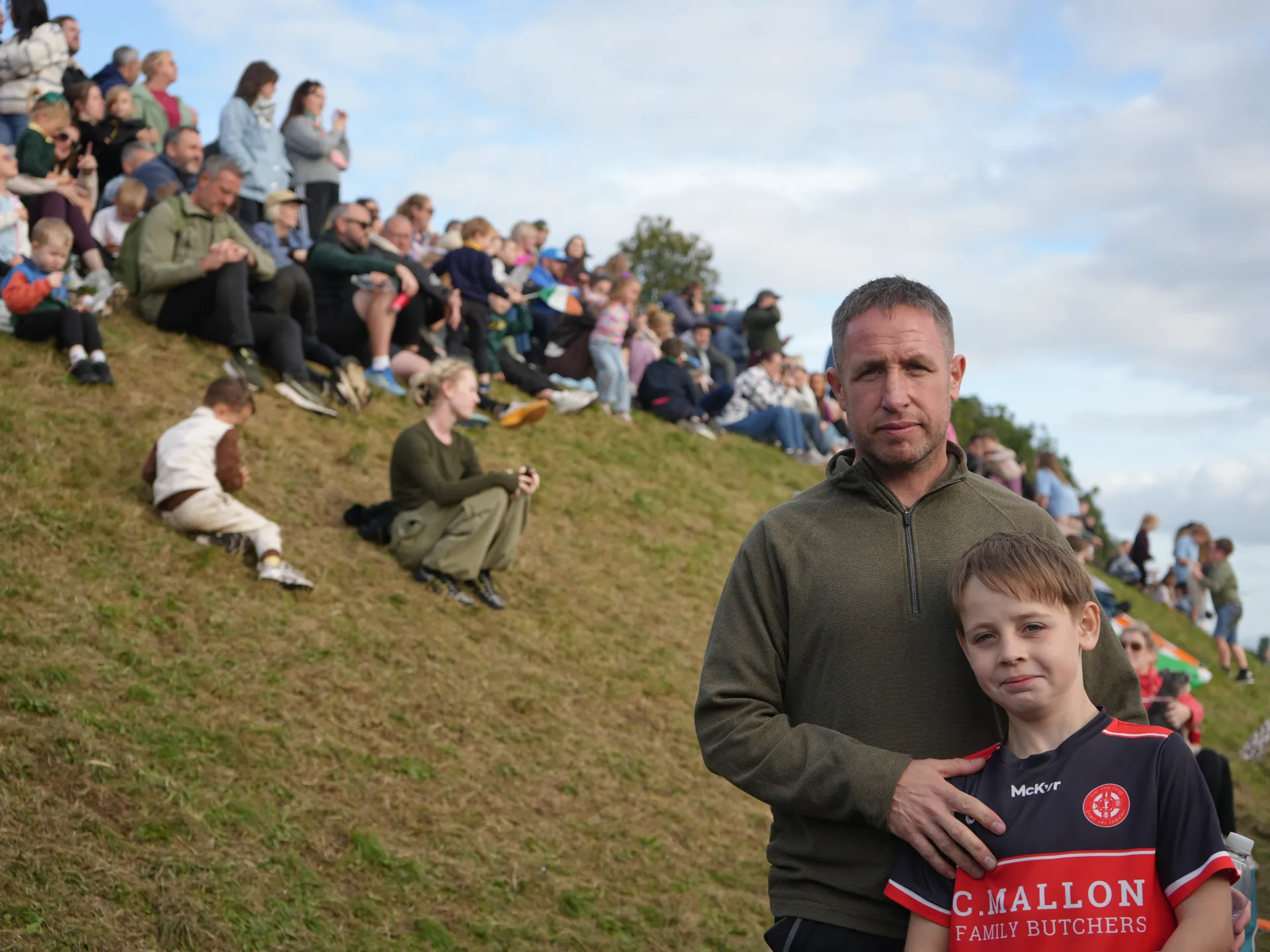 A man and boy in a red and navy football jersey stand before spectators on a grassy slope.