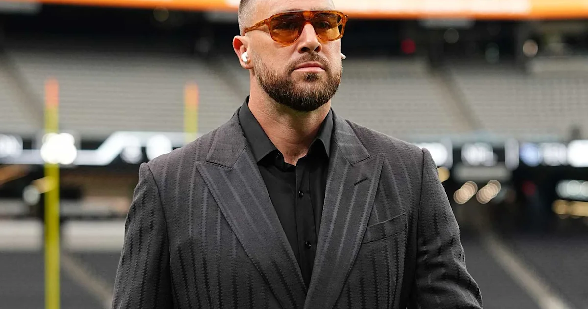 Bearded man in a dark suit, black shirt, and orange-tinted glasses stands in a stadium.