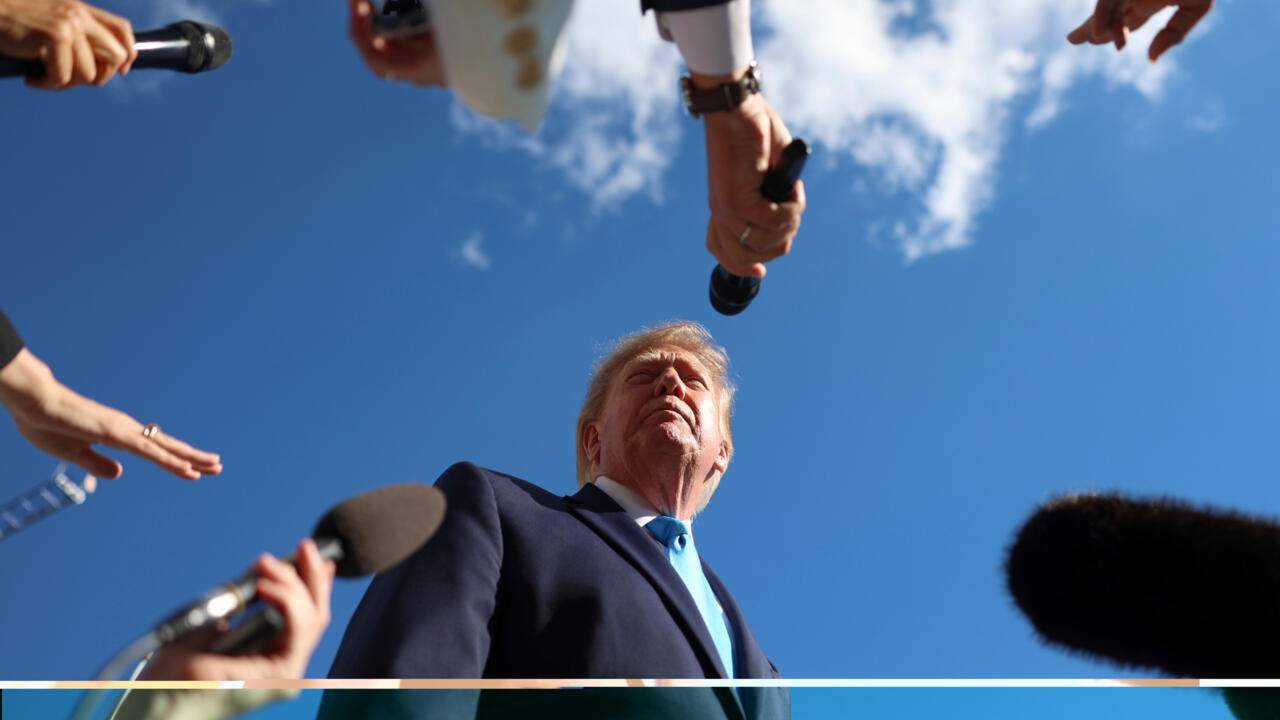 Donald Trump looks upward as reporters extend microphones toward him beneath a blue, cloudy sky.