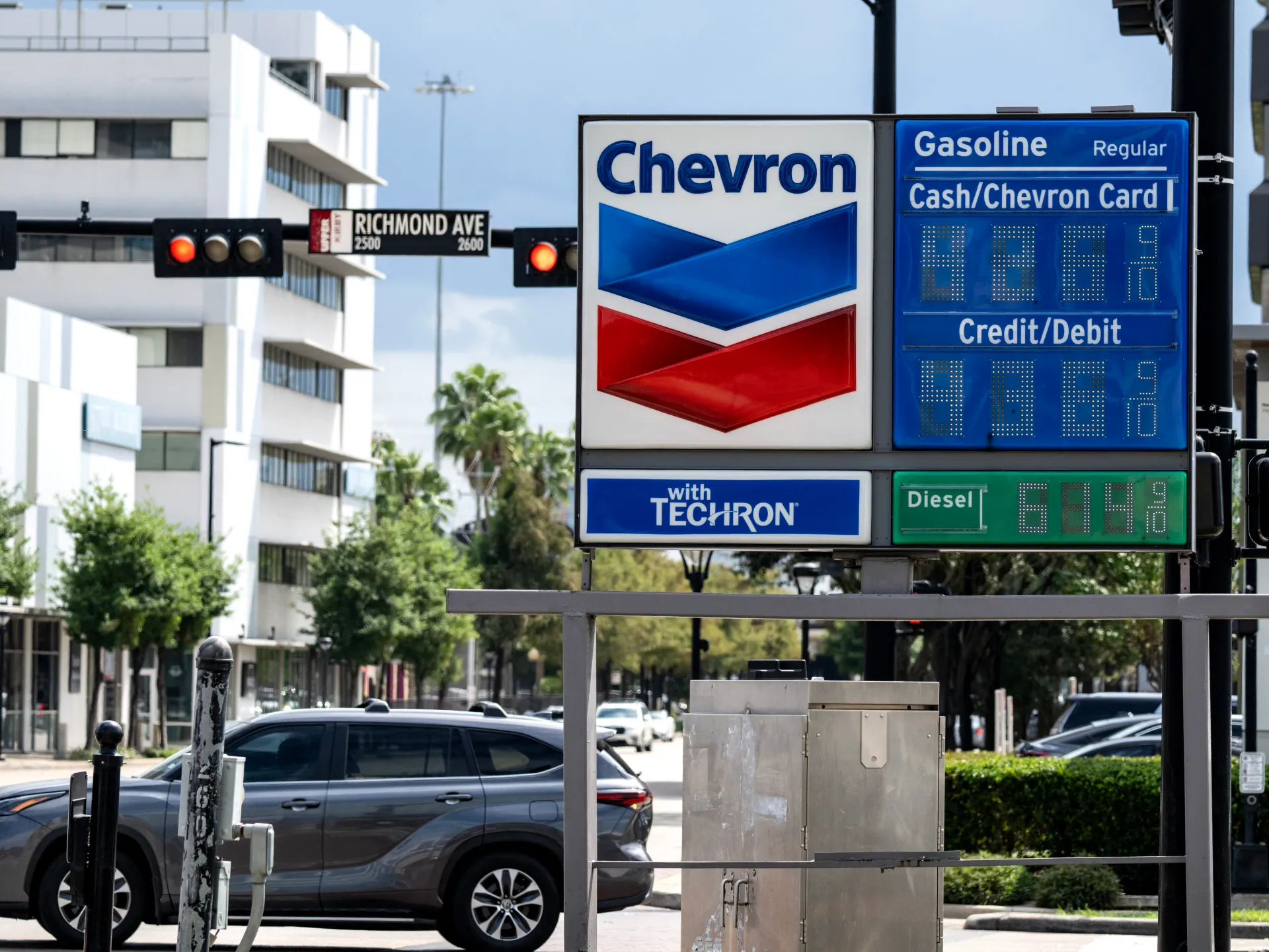 Chevron roadside sign displays gasoline and diesel prices beside a city street with cars and traffic lights.