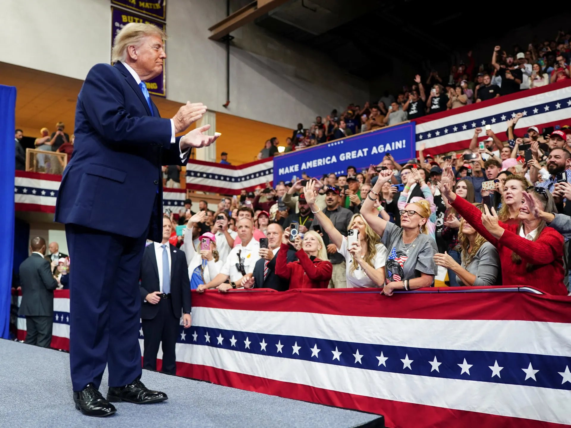 Donald Trump claps onstage as a crowd gathers behind patriotic bunting and a campaign sign.