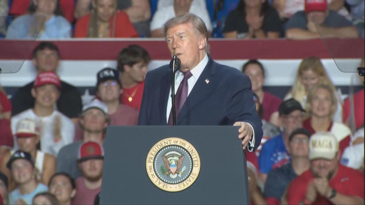Donald Trump speaks at a podium bearing the presidential seal, with a crowd behind him.