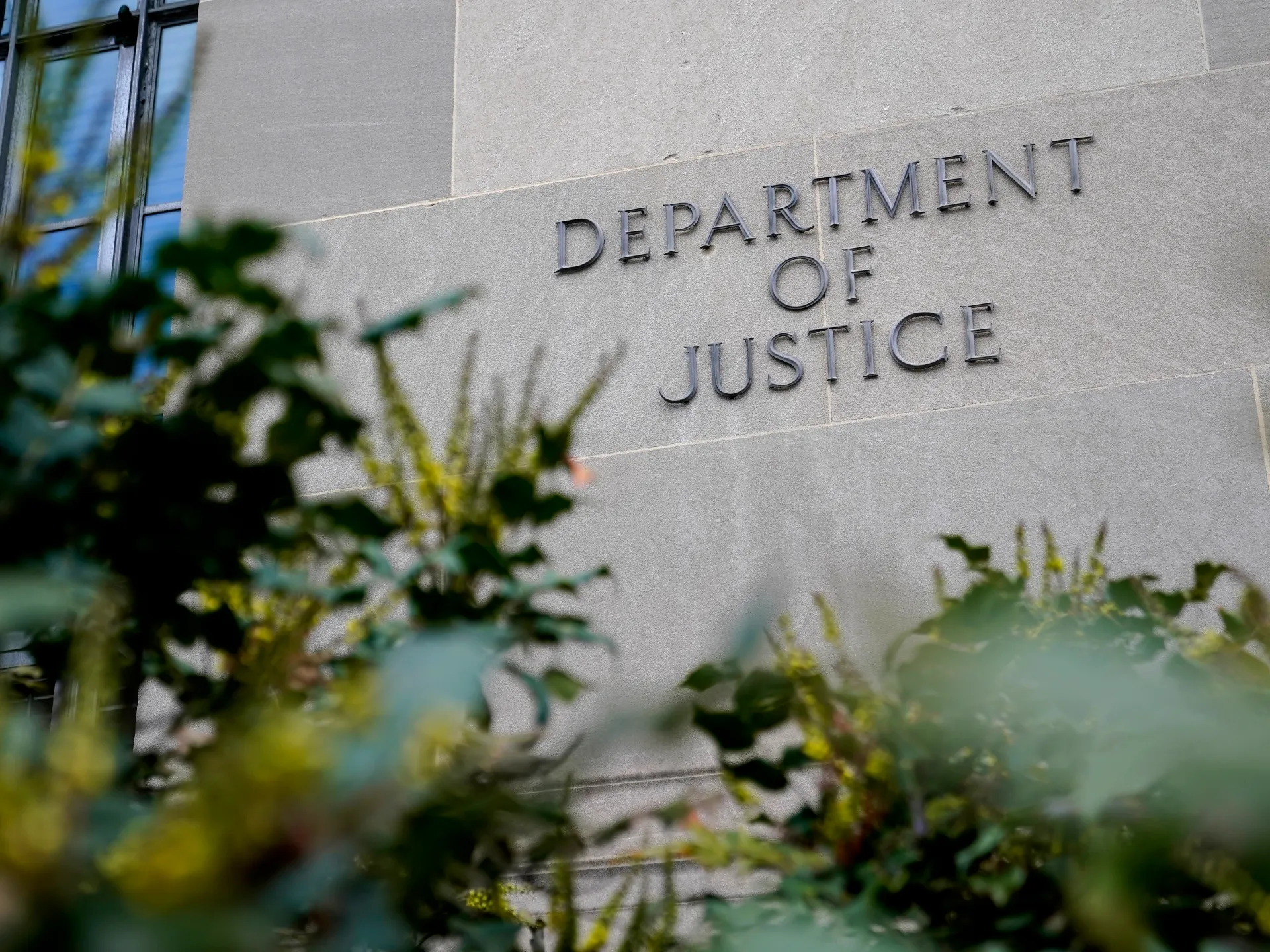Department of Justice lettering on a stone building facade, seen through out-of-focus greenery.