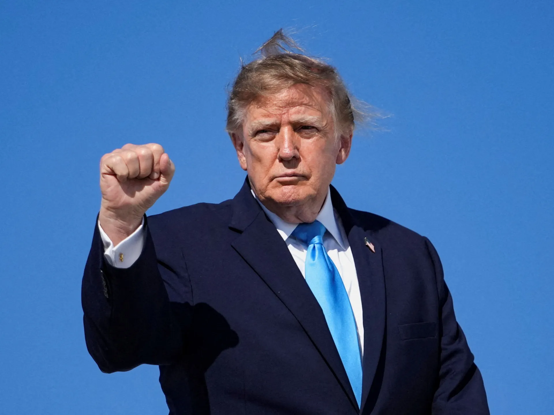 Donald Trump raises a clenched fist while wearing a dark suit and blue tie against a clear blue sky.