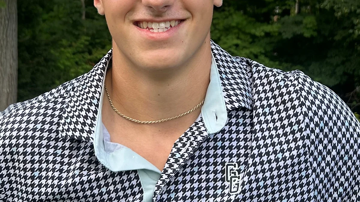 Cropped portrait of a smiling person wearing a black-and-white patterned shirt and gold chain outdoors.